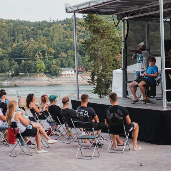 Publikum na skládacích židlích sleduje pódium u vody s barberským křeslem