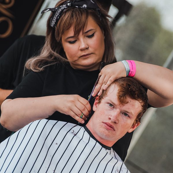 Barberka v šátku stříhá nůžkami zrzavého zákazníka v pruhované pláštěnce