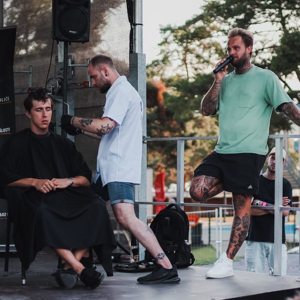 Barber stříhá strojkem zákazníka v černé pláštěnce, lektor vedle komentuje do mikrofonu