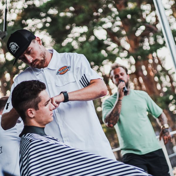 Barber v bílé pracovní košili zastřihuje mladíka v pruhované pláštěnce na pódiu