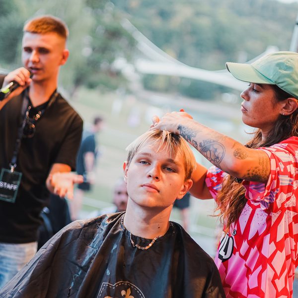 Barberka v zelené kšiltovce upravuje blond vlasy mladíka, vedle moderátor s mikrofonem