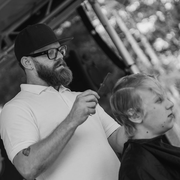 Barber v kšiltovce a brýlích stříhá nůžkami blond zákazníka v pláštěnce