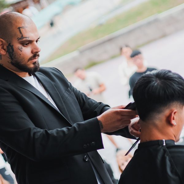Barber s tetováním na obličeji stříhá strojkem tmavovlasého zákazníka