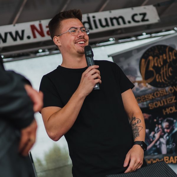Moderátor v brýlích s mikrofonem před bannerem Barber Battle