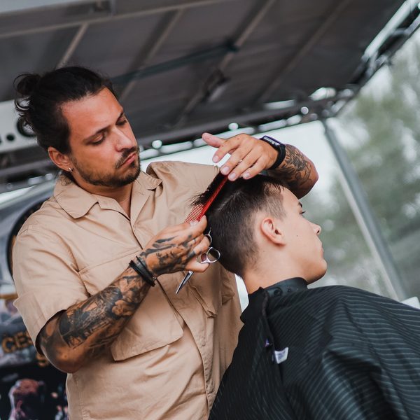 Barber v béžové košili stříhá nůžkami přes hřeben zákazníka v pláštěnce