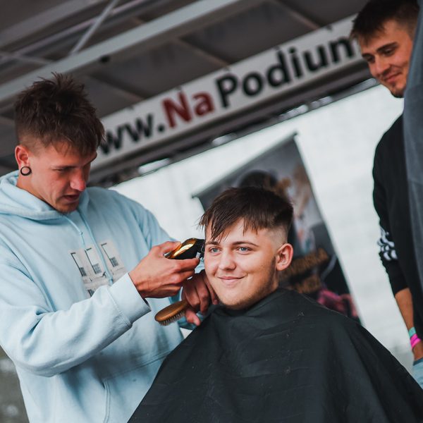Barber v mikině holí strojkem kotlety usmívajícího se mladíka pod bannerem
